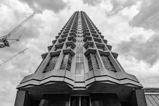 A low angle, monochrome image of a tall building under construction with cranes against a cloudy sky.
