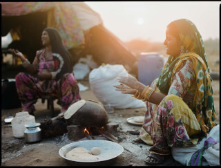 Two women in traditional Indian clothing cooking outdoors over an open fire in a rural setting.