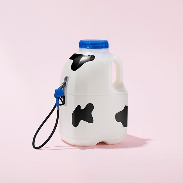 A milk jug with cow print design and a fuel pump nozzle attached on a pink background.