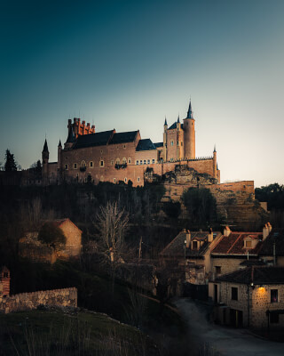 A scenic view of the Alcazar of Segovia, a historic castle in Spain, set against a serene blue sky at dusk.