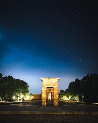 The Debod Temple in Madrid, Spain, stands illuminated against the night sky, surrounded by trees and paving.
