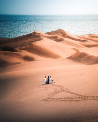 A small group walks across vast sand dunes near the ocean on a bright, sunny day, enjoying the beautiful view.