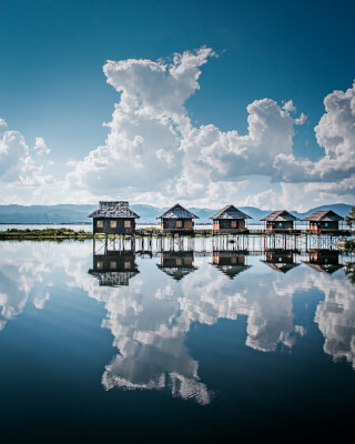 Stilt houses reflect in a calm lake under a blue sky with puffy white clouds in a serene landscape.