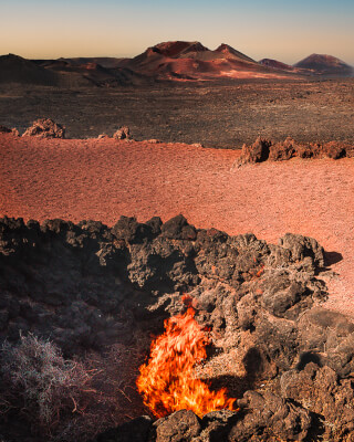 A dramatic volcanic landscape featuring a fiery opening in the earth with a backdrop of mountains and textured terrain.
