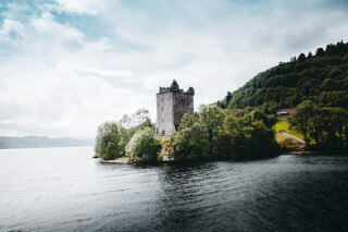 Urquhart Castle sits on the edge of Loch Ness in Scotland, with surrounding trees and water, under a cloudy sky.