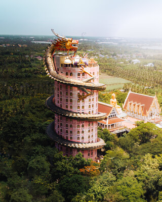 A stunning dragon wraps around a pink Buddhist temple tower in Thailand, surrounded by lush green trees.