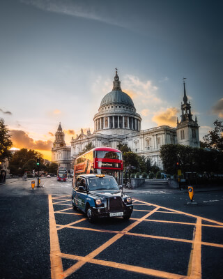 A London taxi and double decker bus pass St Paul's Cathedral on a city street at sunset.