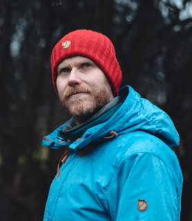 A bearded man in a red beanie and blue jacket looks off-camera in a natural, outdoor setting.