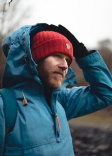 A bearded man wears a blue jacket and red beanie while gazing thoughtfully outdoors.