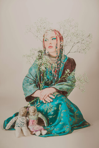 A woman with braided hair sits adorned with flowers, holding baby's breath and dolls, radiating serene vintage style.