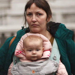 A woman carries a baby in an ergobaby carrier outdoors, both looking at the camera.