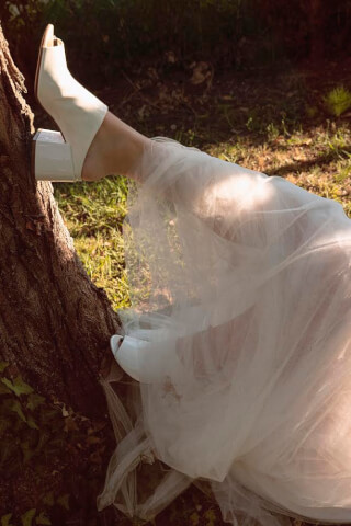A person's legs in white open-toe heels and a flowing dress rest against a tree in a dreamy outdoor setting.