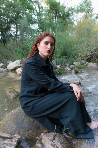 A woman in a dark outfit sits on a rock in a river surrounded by trees.