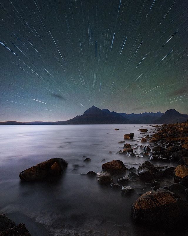 Star trails over a rocky coastline and calm sea, with mountains silhouetted against the night sky, creating a serene seascape.